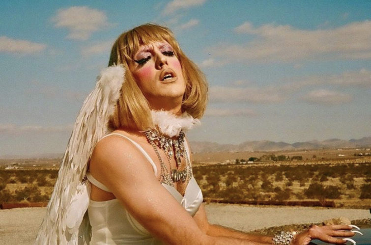 A close-up of the winged woman details her extravagent necklaces, excessively long and manicured nails, and a face made-up with pink blush, winged eyelashes, and glittery-purple eyeshadow.